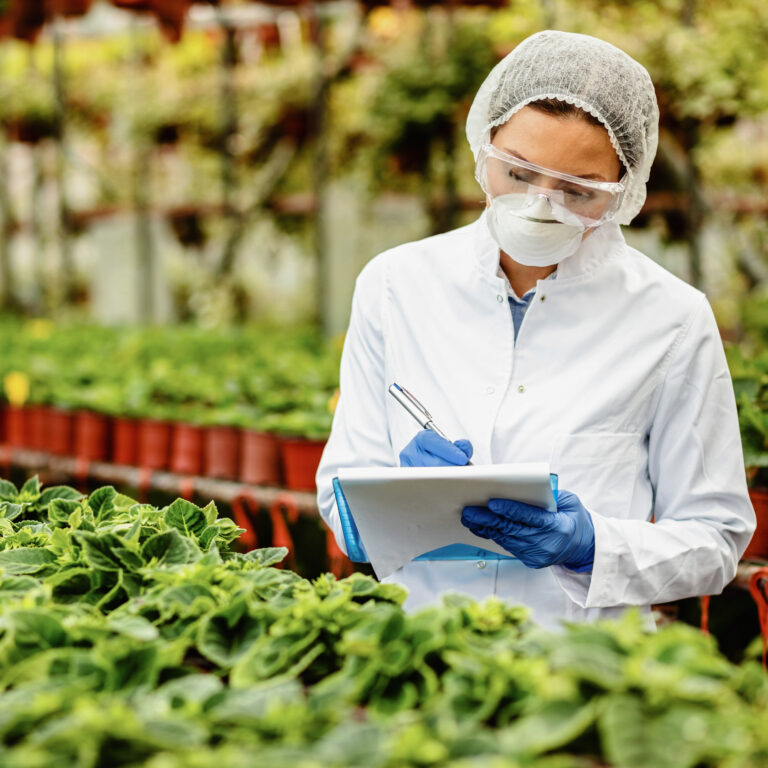 Female biotechnologist inspecting potted plants in plant nursery and writing notes into clipboard.